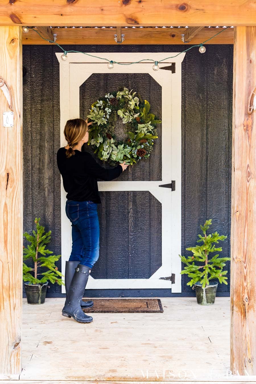 woman hanging wreath on black farmhouse exterior with white trim and holiday decor