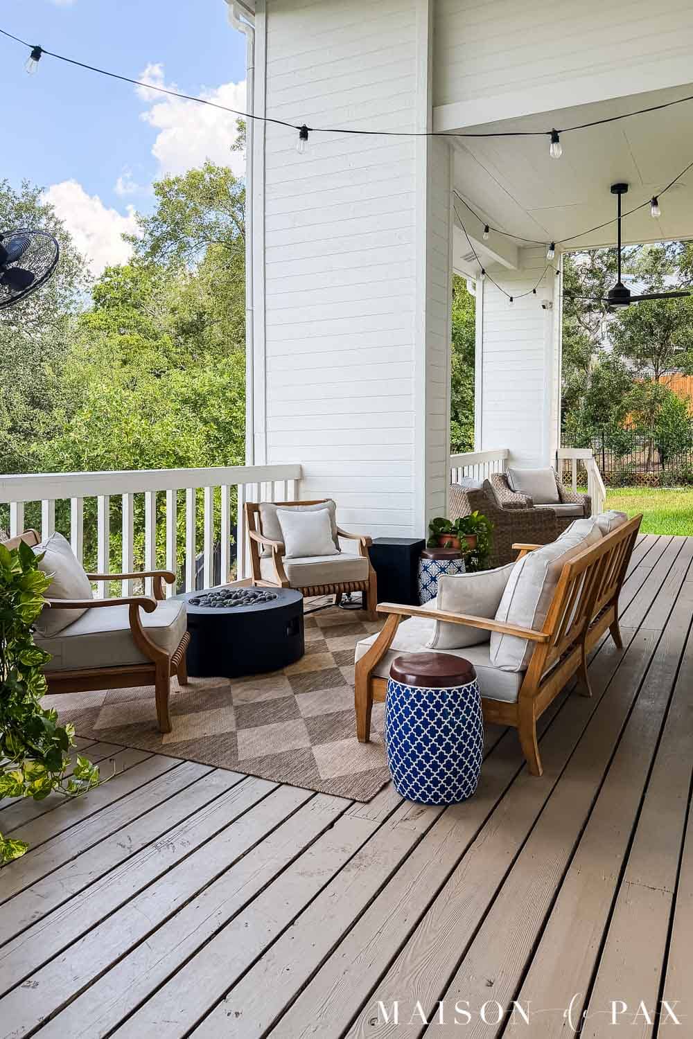 Covered wood porch approximately 14 months after staining, with warm beige deck boards, teak seating, outdoor rug, string lights, and no visible peeling from standing distance.