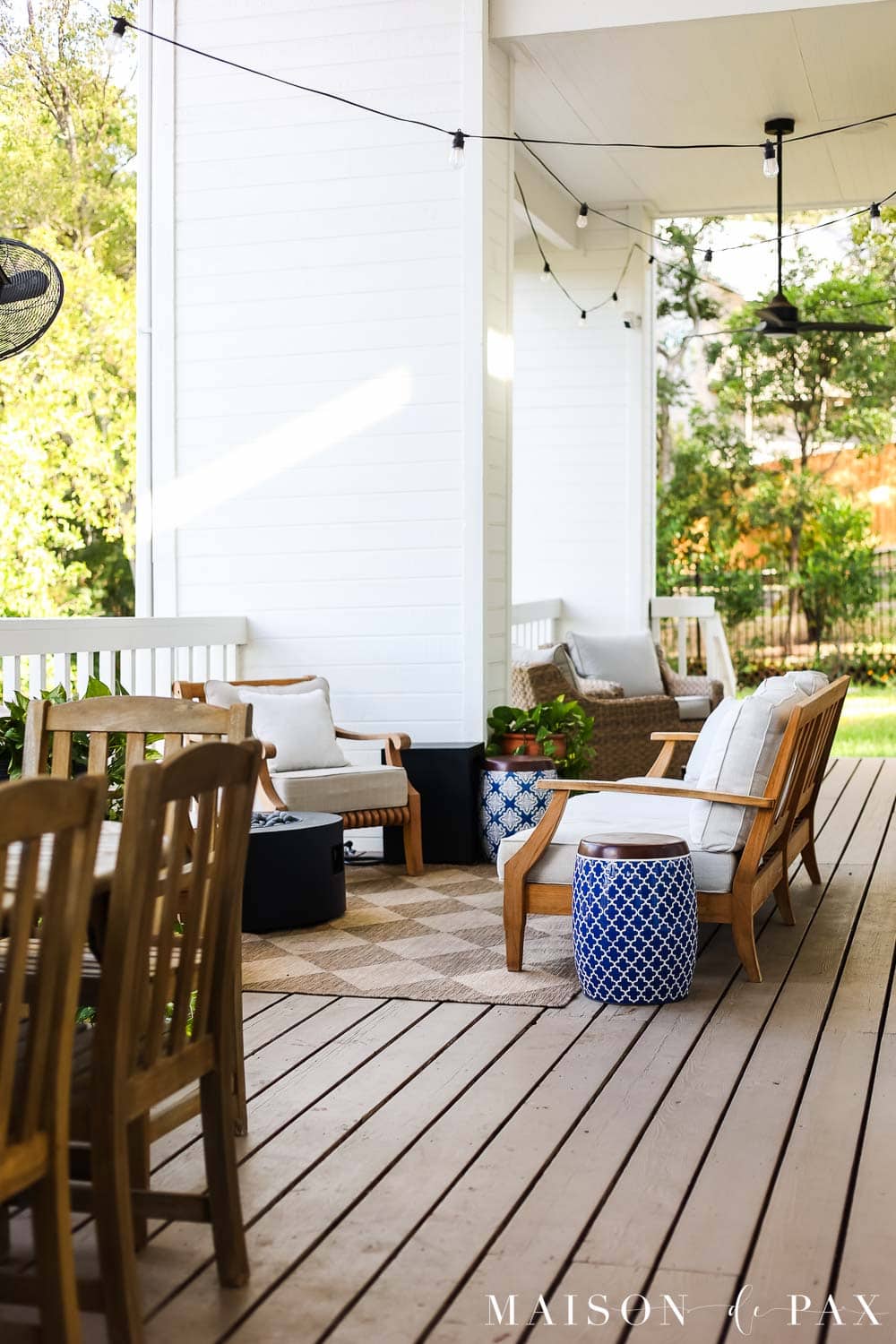 Finished covered porch with warm beige solid-stained wood decking, white exterior walls, black doors, teak seating, string lights, and neutral outdoor cushions.