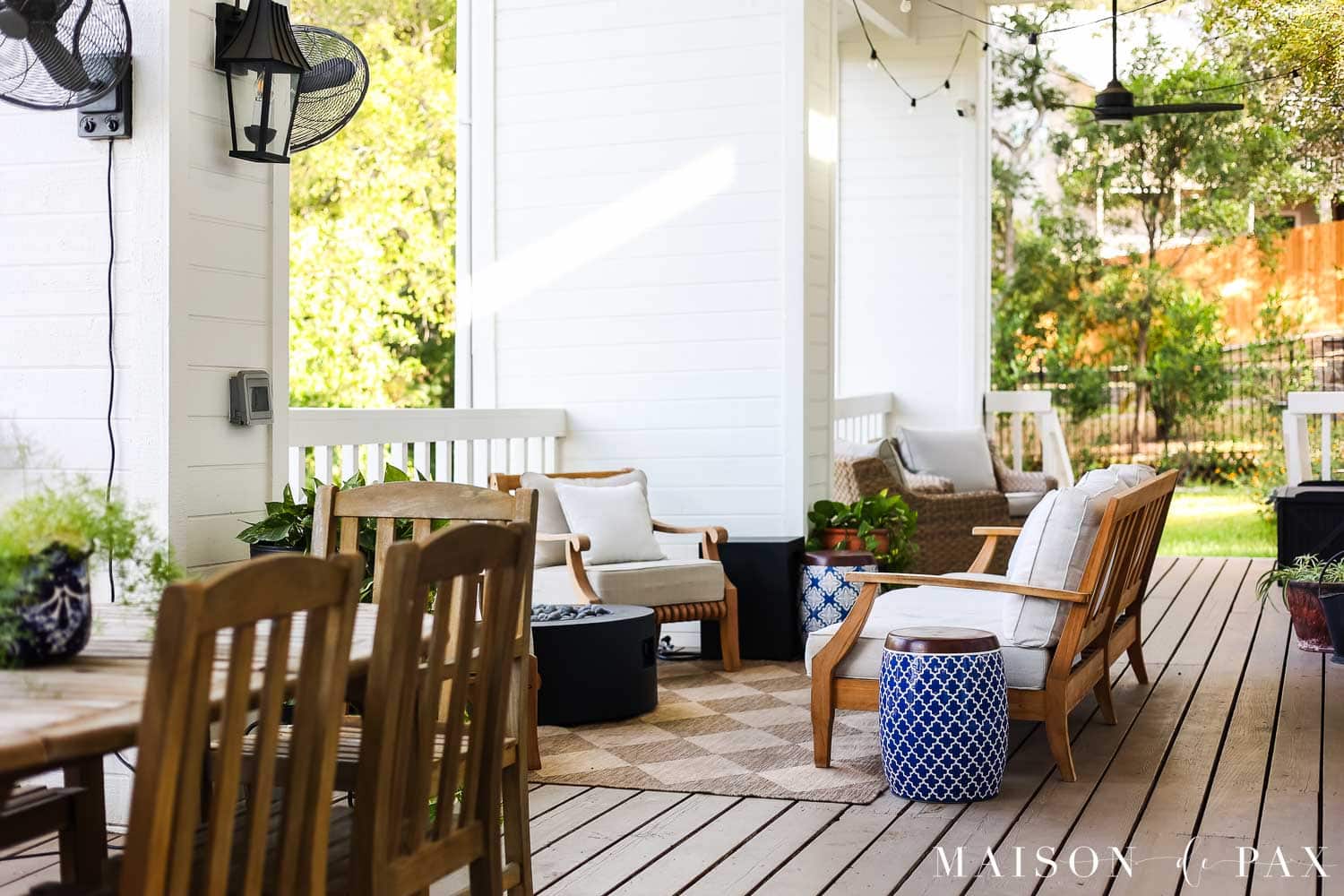 Finished covered porch with warm beige solid-stained wood decking, white exterior walls, black doors, teak seating, string lights, and neutral outdoor cushions.