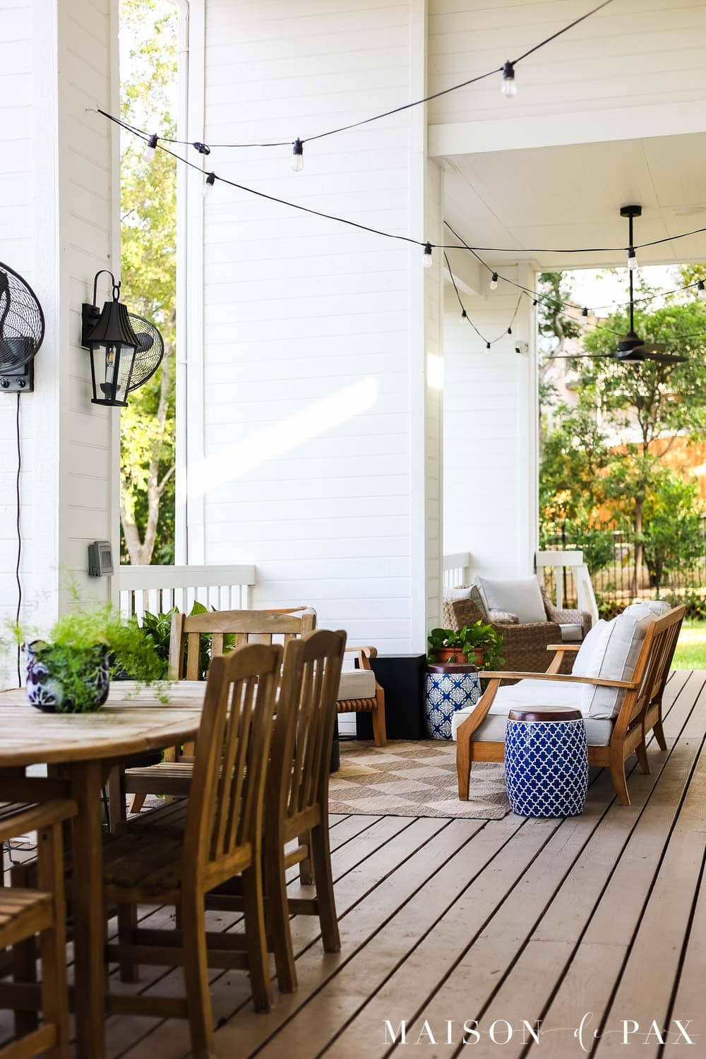 Covered white porch with warm neutral stained deck boards, natural teak outdoor furniture, neutral cushions, and black exterior accents.