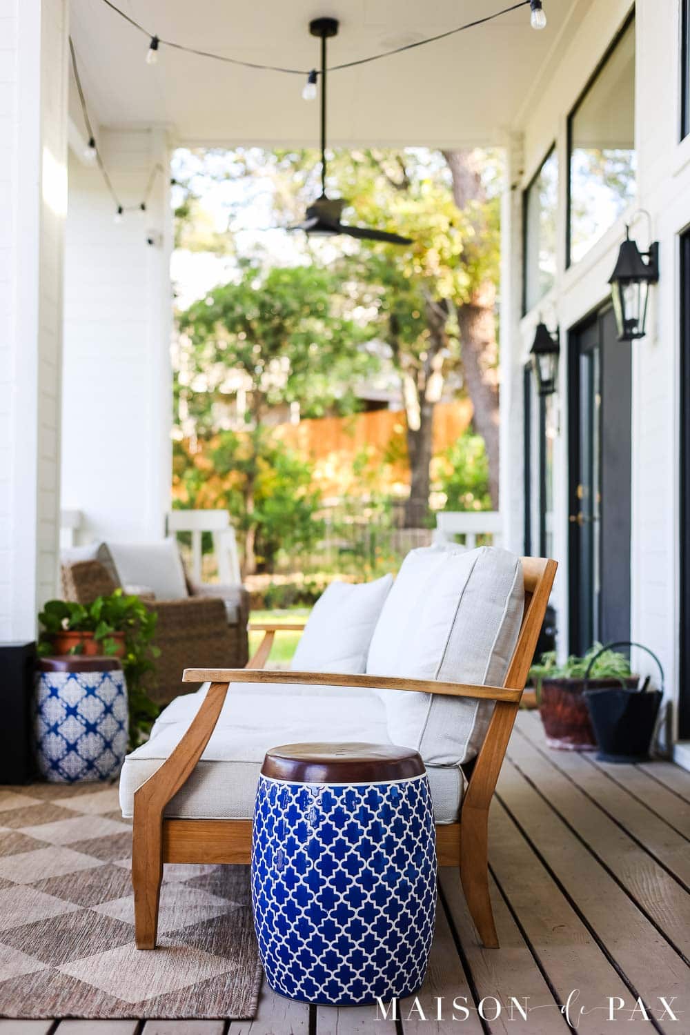 Finished outdoor living area on a covered porch with warm beige deck boards, teak furniture, neutral textiles, and blue accents.