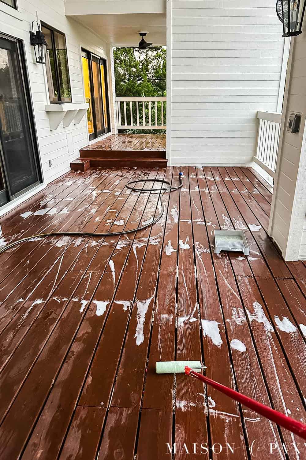 Reddish-brown wood deck covered with Cabot Wood Cleaner during washing, with a roller, hose, and cleaning solution visible.